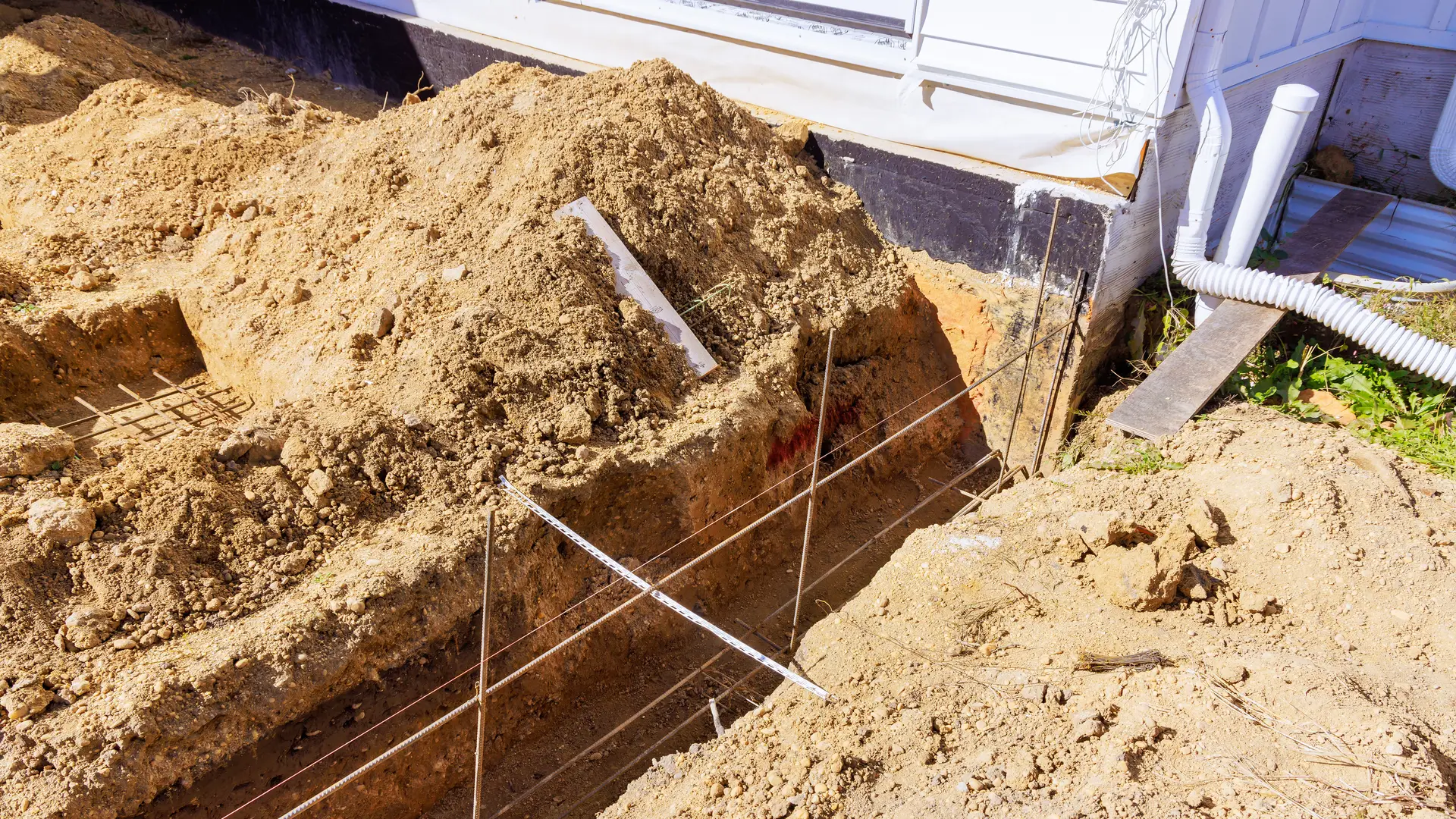 Trench dug beside a house for drainage, with white downspout pipes