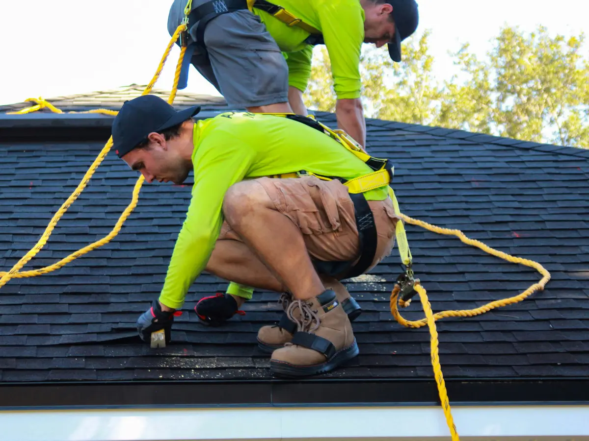 Two roofers in safety harnesses working on a dark shingle roof