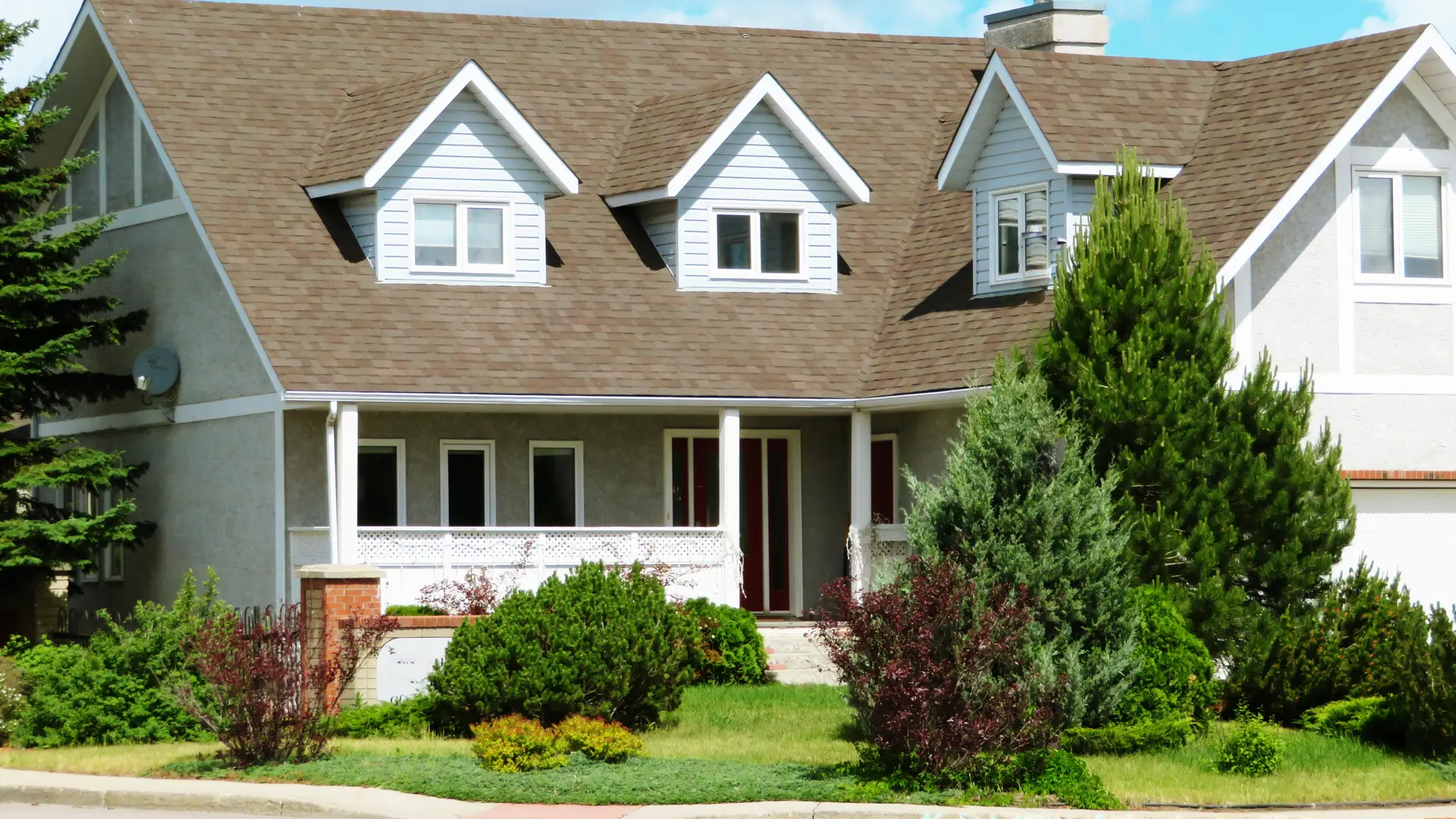 House with pale siding, dormers and a brown shingle roof behind a lawn
